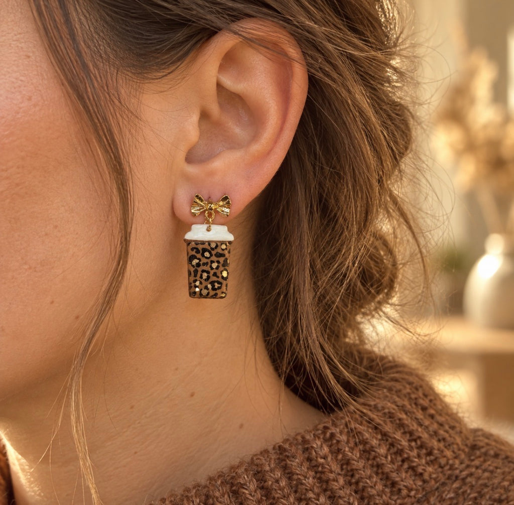 A close-up of a person's ear wearing a dangling earring. The earring features a small gold bow on a white and leopard print takeaway coffee cup.