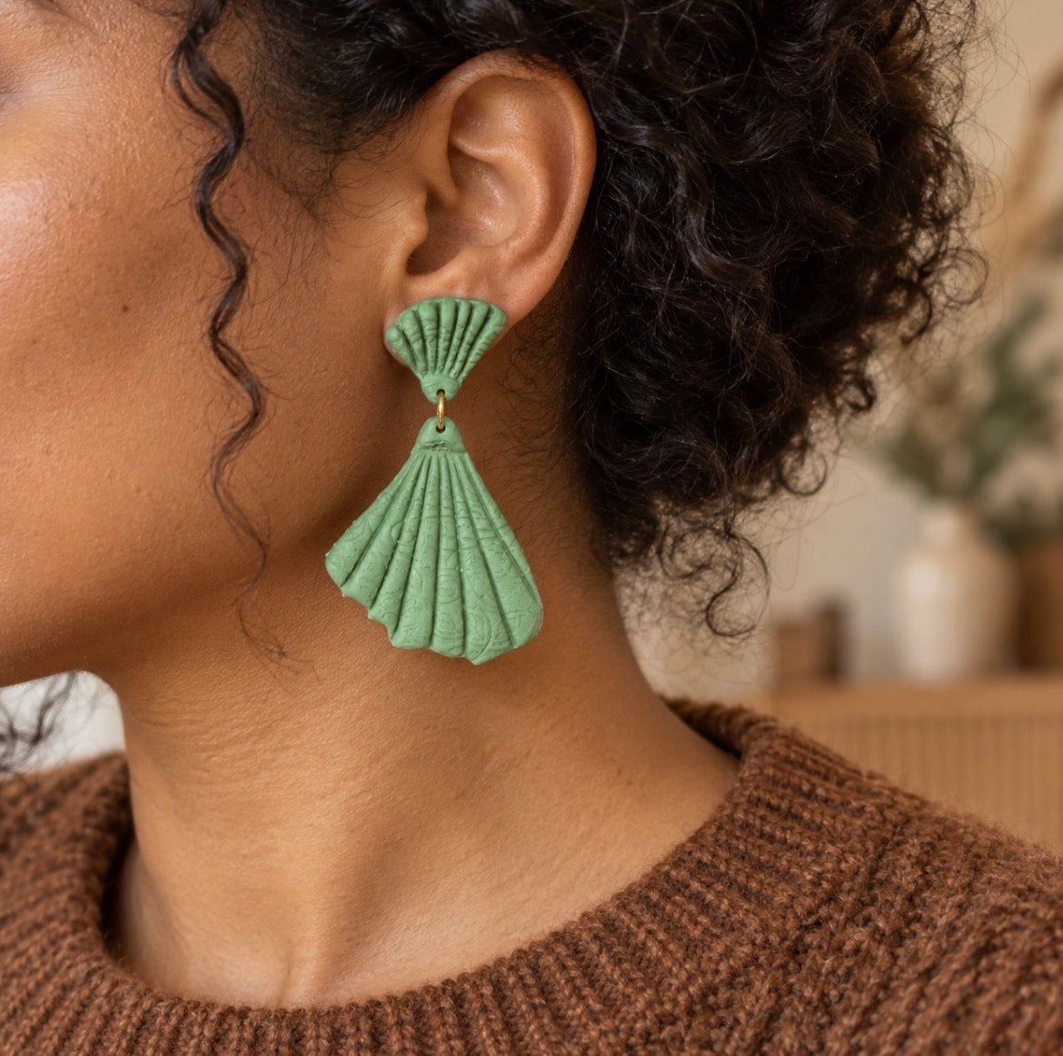 A close-up side profile of a person wearing a textured sage green dangle earring with a scalloped fan design and a small gold-tone connector, paired with a brown knit sweater.