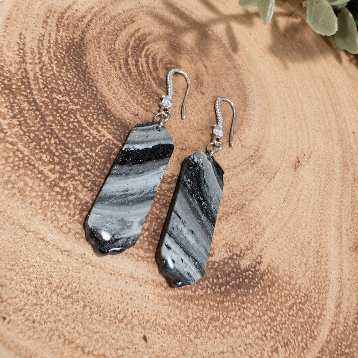 A pair of dangle earrings with polished black and white striped stone slices, hanging from silver fishhook wires, displayed on a rustic wood slice background with green leaves in the corner.