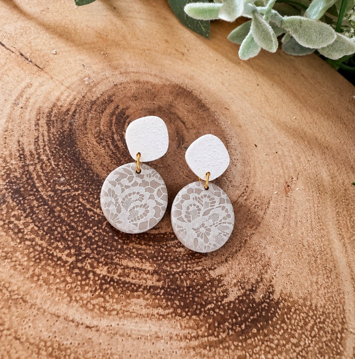 A pair of handmade dangle earrings with white circular floral lace discs and white square studs, resting on a rustic wood slice background with yellow flowers.