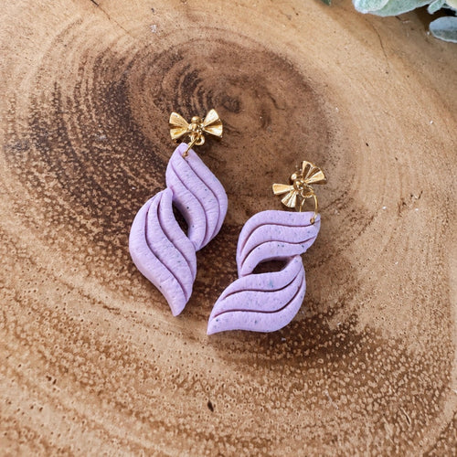 A pair of blush pink polymer clay dangle earrings with gold-tone bow stud backs, displayed on a rustic wood slice background