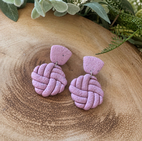 A pair of handmade lavender polymer clay dangle earrings with a knotted ball design and a small fan-shaped top, displayed on a rustic wooden slice with green foliage in the background.