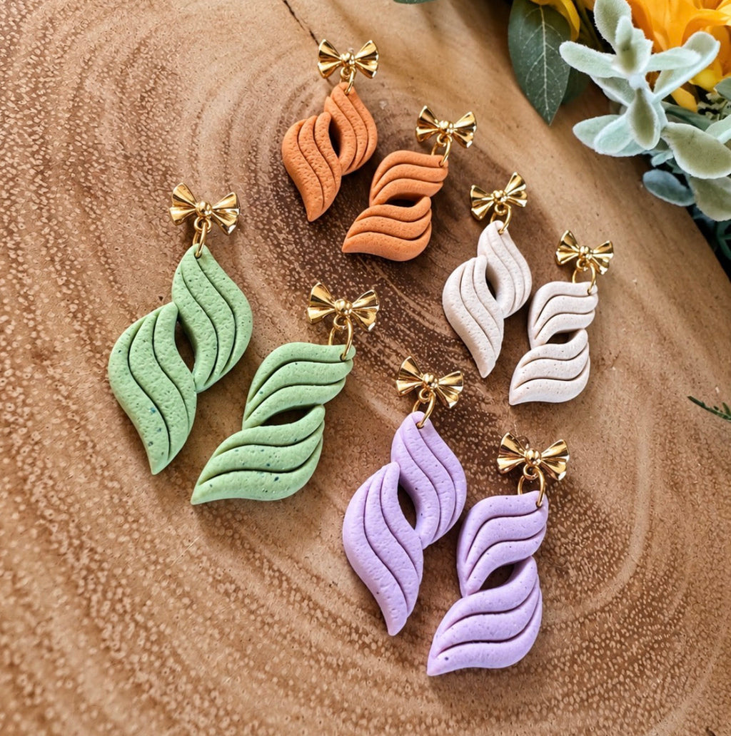 Four pairs of sculptural, leaf-shaped dangle earrings in pastel green, orange, white, and lavender, each featuring a gold-tone bow detail at the top, arranged on a textured wood slice background with yellow flowers in the corner.