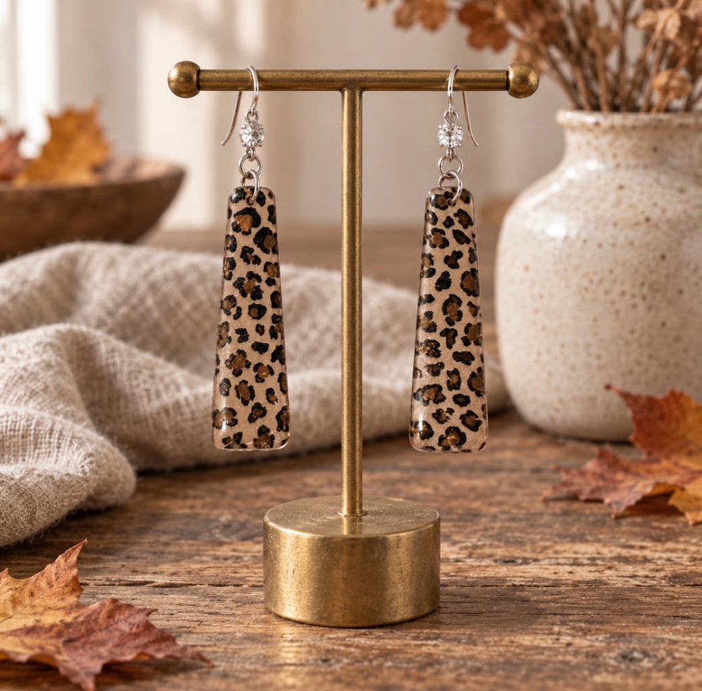 A pair of long rectangular drop earrings with a beige, brown, and black leopard print pattern, displayed on a gold-colored T-stand. The stand is set on a rustic wooden table with a beige linen cloth, a ceramic vase with dried flowers, and scattered autumn leaves in the background.