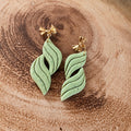 A pair of light green leaf-shaped drop earrings with gold butterfly posts, displayed on a rustic wooden slice background.