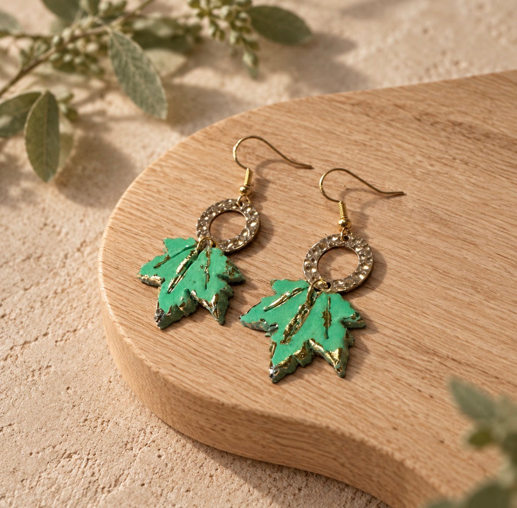 A pair of dangling earrings with green maple leaf charms and gold-tone hooks, displayed on a curved wooden slice against a sandy beige background with soft green leaves.