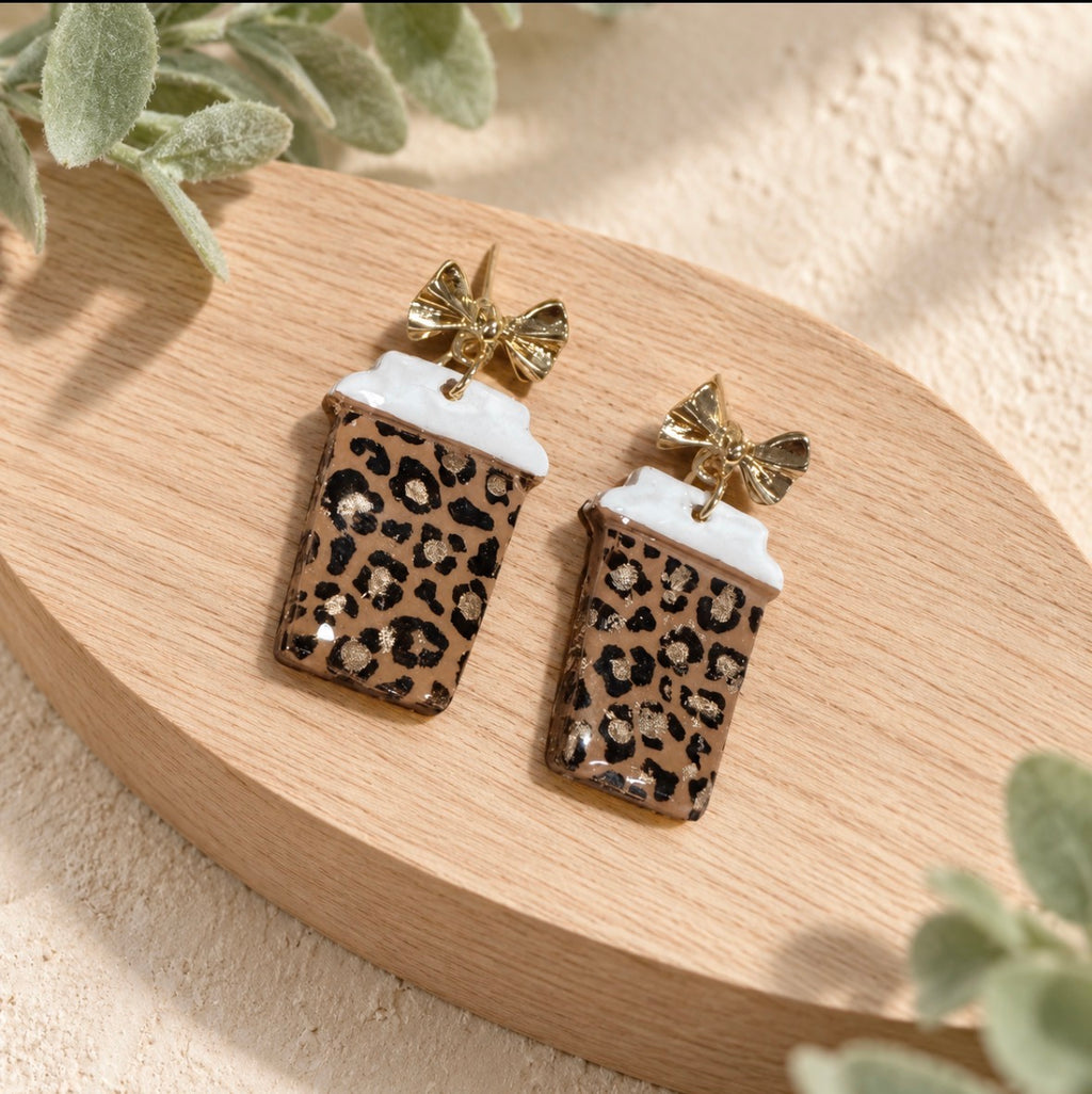 A pair of coffee cup earrings with a brown leopard print pattern, white foam tops, and gold bow details, displayed on a curved wooden tray with soft green leaves.