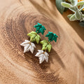 A pair of handmade leaf-shaped earrings with white bottom, light green middle, and dark green top layers, displayed on a wooden surface with yellow flowers in the background.