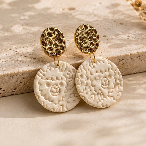 A pair of gold and cream drop earrings with textured circular studs and larger embossed disc pendants with an alpaca pattern, displayed on a beige stone surface.