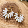 A pair of dangling earrings with white enamel-covered teardrop-shaped charms outlined in gold, hanging from gold-tone fishhook wires, displayed on a textured wood slice background.