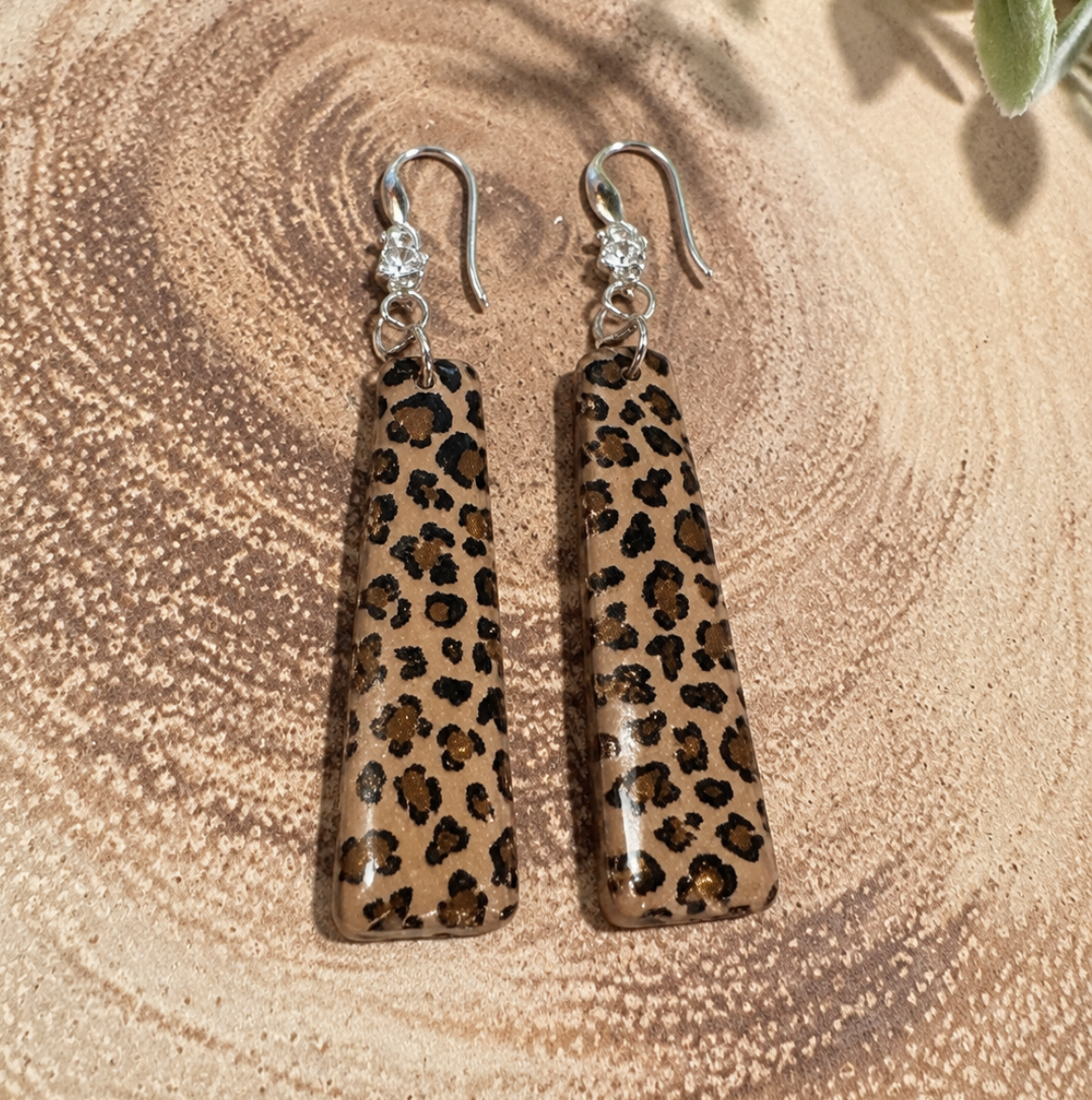A pair of dangling earrings with leopard-print rectangular charms and silver fishhook wires, displayed on a wood slice background inside a photo-editor interface.