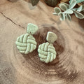 A pair of handmade light sage green clay earrings with a textured braided knot design, displayed on a rustic wood slice background with dried flowers.
