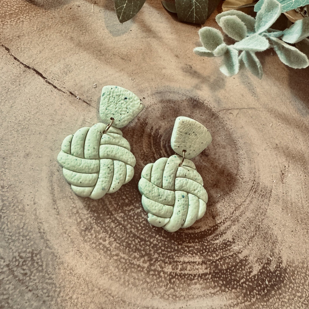 A pair of handmade light sage green clay earrings with a textured braided knot design, displayed on a rustic wood slice background with dried flowers.