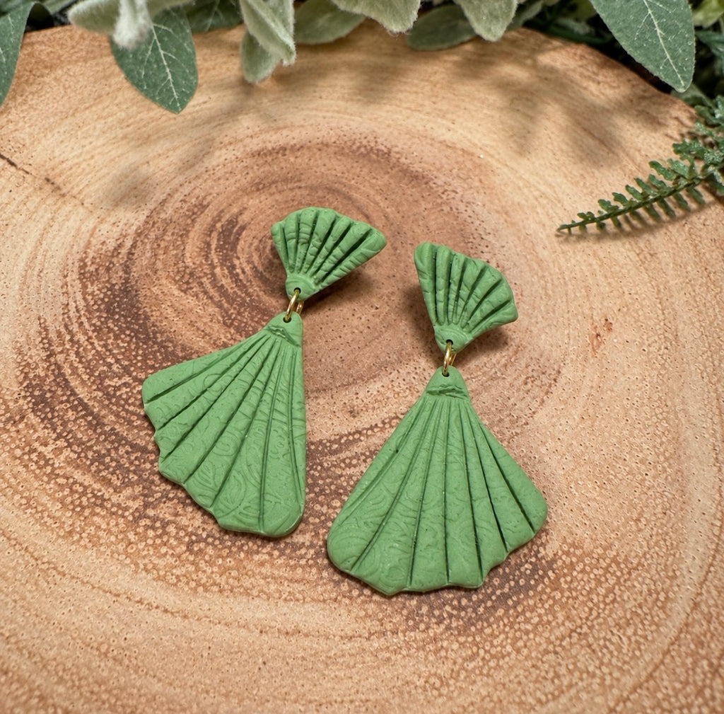 A pair of green shell-shaped dangle earrings with a textured, pleated fan design, displayed on a rustic wooden slice background with green foliage.