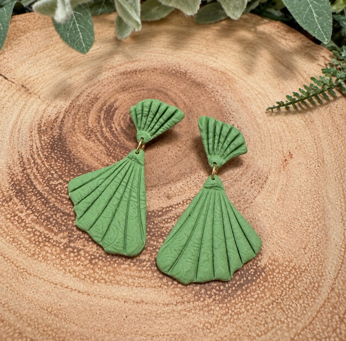 A pair of green shell-shaped dangle earrings with a textured, pleated fan design, displayed on a rustic wooden slice background with green foliage.