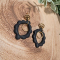 A pair of black and gold statement earrings displayed on a rustic wood slice background with green leaves and yellow flowers.
