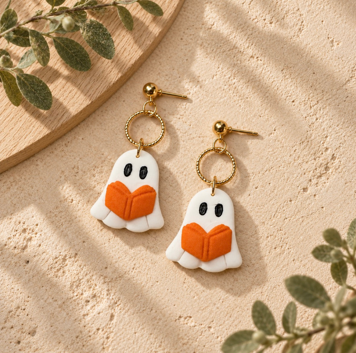 A pair of gold-tone dangle earrings shaped like white ghosts reading orange books, displayed on a textured beige surface with decorative green leaves and a wooden tray.