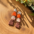 A pair of handmade dangle earrings styled on a textured wooden surface with natural sunlight and shadows. The earrings feature a white ghost, an orange textured ghost, a beige and brown marble ghost, and a brown textured ghost, finished with small gold bow accents at the top.