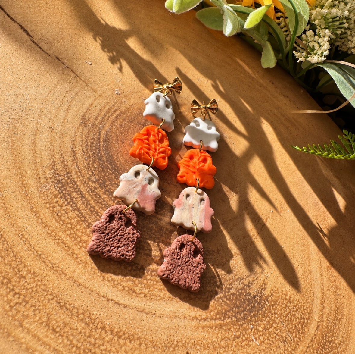 A pair of handmade dangle earrings styled on a textured wooden surface with natural sunlight and shadows. The earrings feature a white ghost, an orange textured ghost, a beige and brown marble ghost, and a brown textured ghost, finished with small gold bow accents at the top.