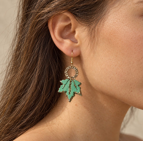 A close-up of a woman's ear wearing a dangling earring with a gold-tone circular ring and a green maple leaf charm outlined in gold.