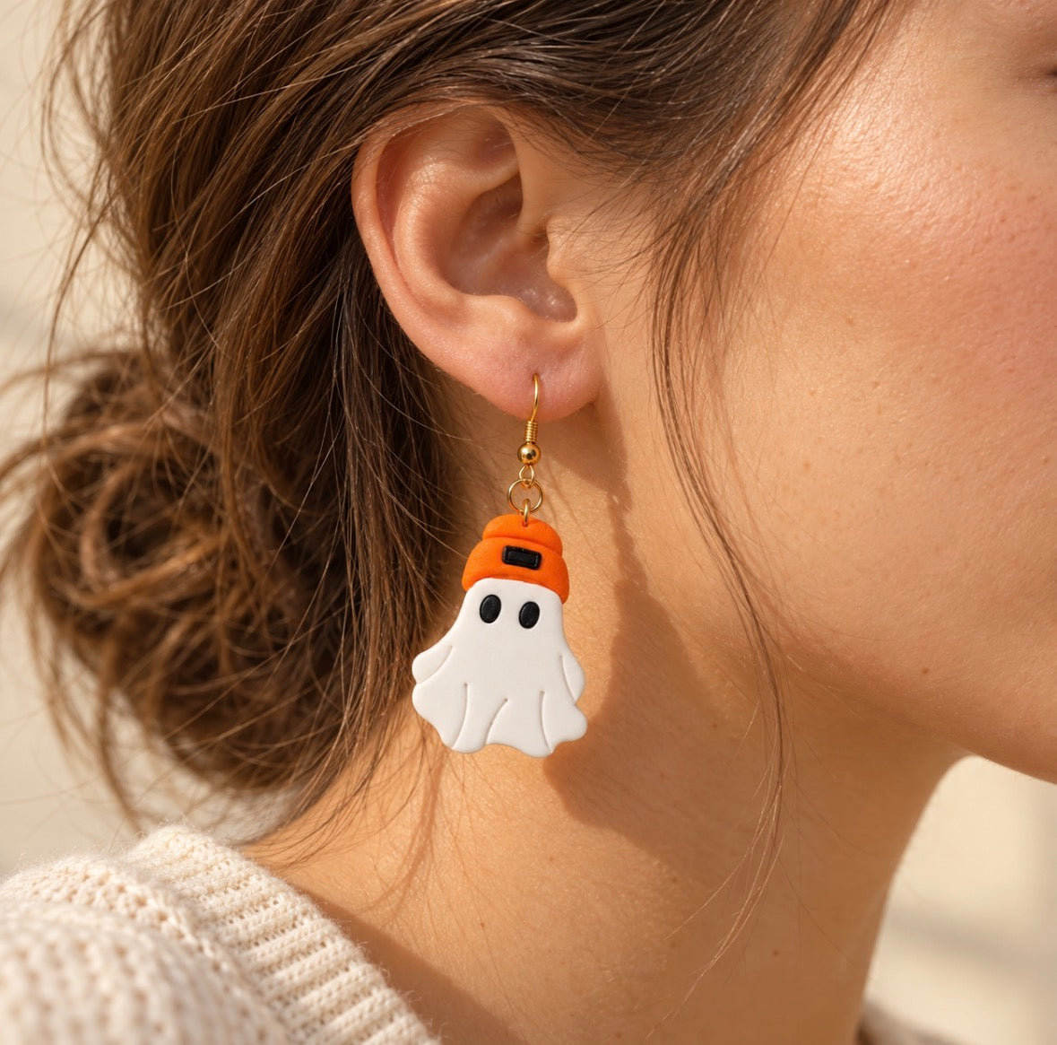 A close-up of a person's ear wearing a dangling ghost earring. The ghost charm is white with black eyes and wears an orange hat, suspended from a gold hook.