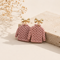 A pair of miniature pink knit sweater earrings with gold bow stud tops, displayed on a beige stone block with dried flowers in the background.