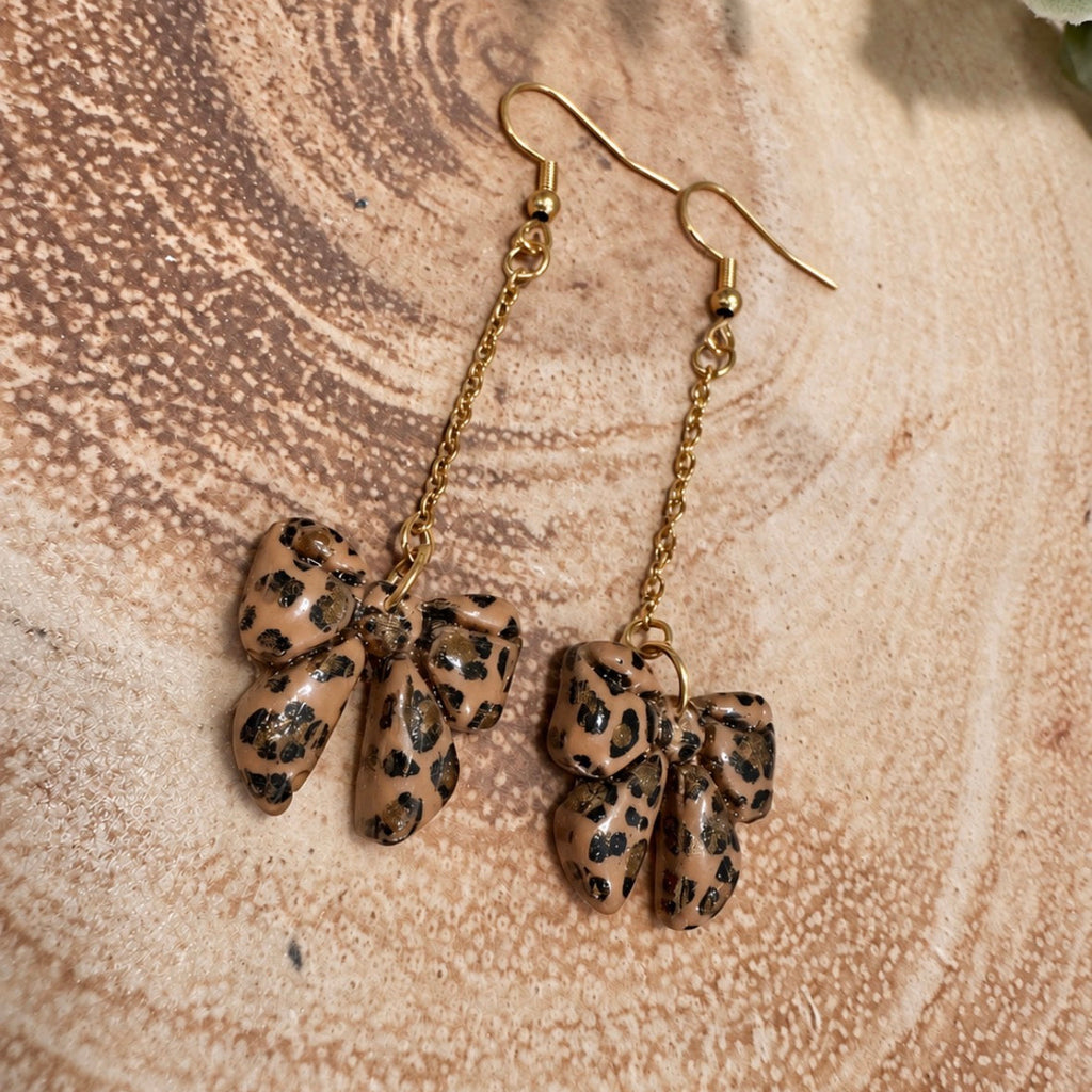 A pair of dangling earrings with gold-tone fishhook wires and chains, each ending in a small leopard-print bow charm, displayed on a textured wood slice background.