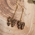 A pair of dangling earrings with gold-tone fishhook wires and chains, each ending in a small leopard-print bow charm, displayed on a textured wood slice background.
