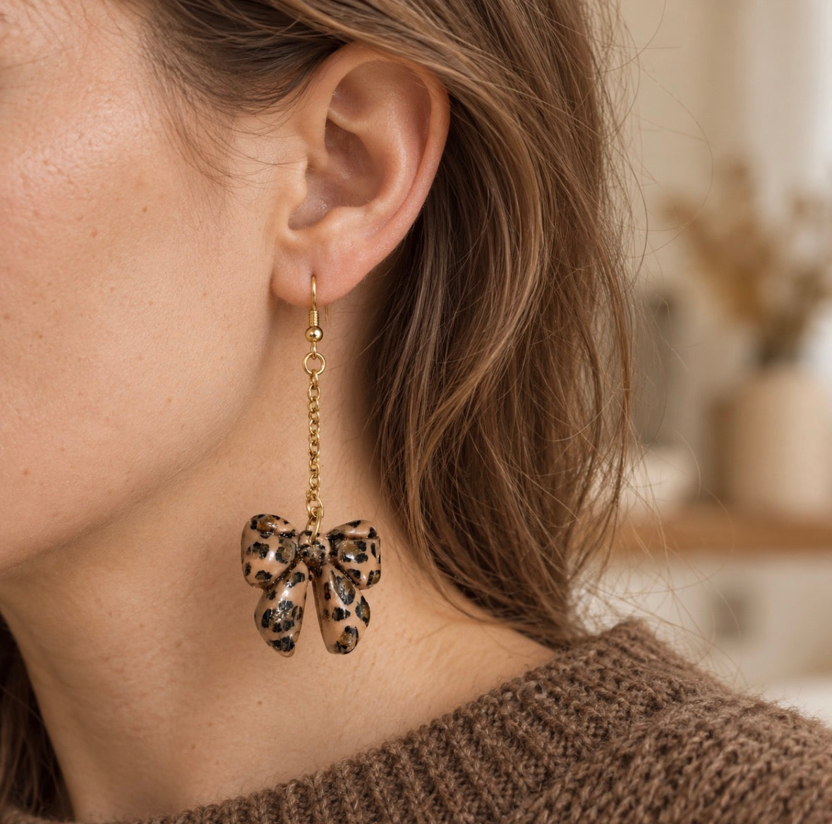 A close-up of a woman's ear wearing a dangling gold-tone earring with a leopard-print bow charm, against a softly blurred indoor background.